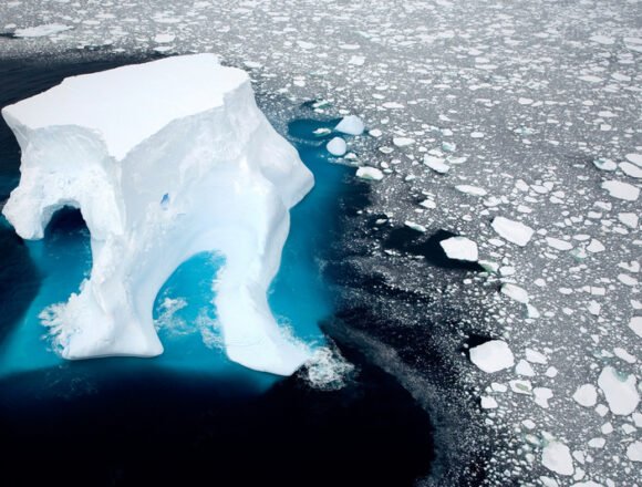 Alerta en la Antártida: liberación de metano desde el subsuelo marino Vista aérea de un iceberg gigante emergiendo del océano en la Antártida, rodeado por fragmentos de hielo flotante y aguas oscuras, símbolo del deshielo polar.