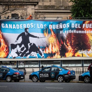 Cartel de Greenpeace frente a una avenida en Buenos Aires, donde se lee "Ganaderos: los dueños del fuego", acompañado de imágenes de vacas entre llamas y una figura señalando, con patrullas de la Policía de la Ciudad estacionadas abajo.