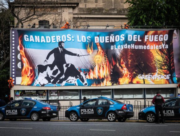 Greenpeace exige ley contra ecocidios en Argentina Cartel de Greenpeace frente a una avenida en Buenos Aires, donde se lee "Ganaderos: los dueños del fuego", acompañado de imágenes de vacas entre llamas y una figura señalando, con patrullas de la Policía de la Ciudad estacionadas abajo.