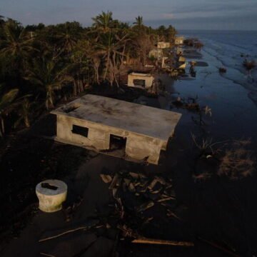 Casas destruidas y parcialmente sumergidas por la erosión costera en una zona tropical, con el mar avanzando sobre antiguas construcciones de cemento en ruinas, rodeadas de palmeras y árboles afectados por el agua salada.