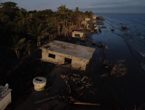 Casas destruidas y parcialmente sumergidas por la erosión costera en una zona tropical, con el mar avanzando sobre antiguas construcciones de cemento en ruinas, rodeadas de palmeras y árboles afectados por el agua salada.