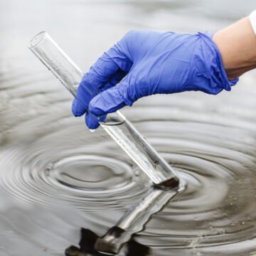 Investigador con guantes de látex azul toma una muestra de agua con un tubo de ensayo en un cuerpo de agua posiblemente contaminado.