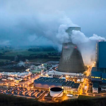 Vista aérea de una planta industrial con una gran torre de enfriamiento expulsando vapor al cielo, rodeada por instalaciones iluminadas. Se observa un paisaje rural bajo un cielo nublado.