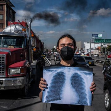 Hombre con barbijo sostiene una radiografía de pulmones frente a una calle con tráfico pesado y humo negro saliendo de un camión, simbolizando la contaminación del aire.