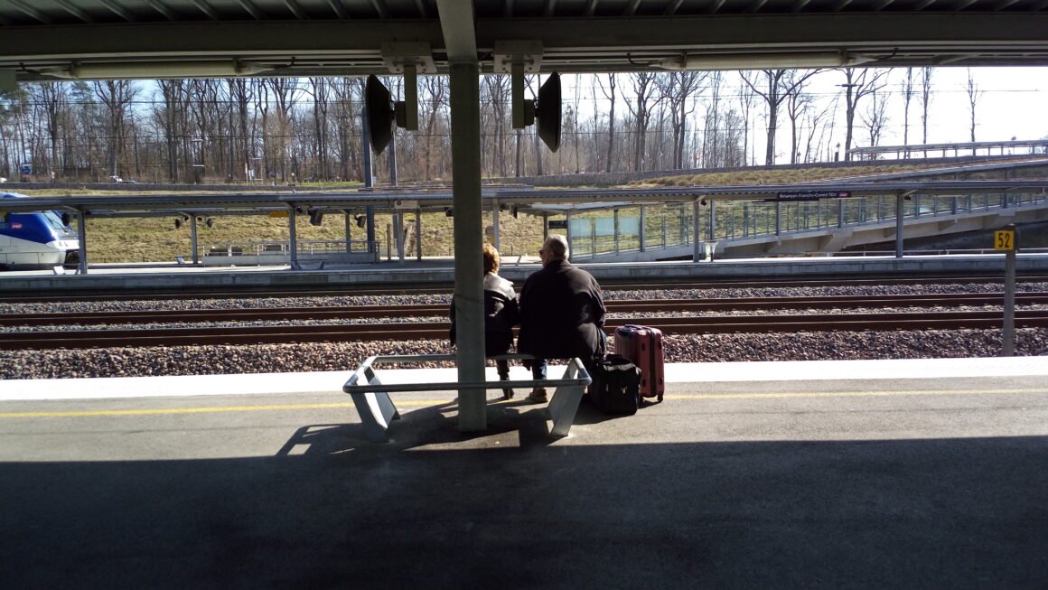Dos personas mayores sentadas en un banco de una estación de tren, con maletas, mirando las vías, en una escena tranquila pero cargada de historia e ingeniería ambiental.