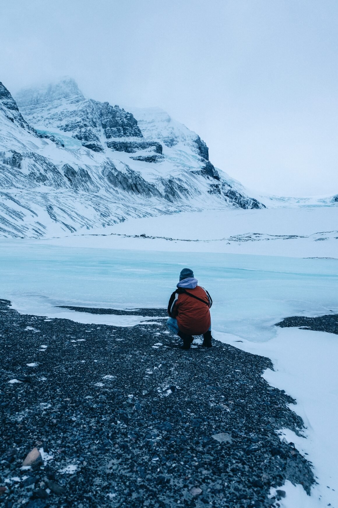 Persona sentada frente a un glaciar en retroceso, rodeada de nieve y montañas heladas.