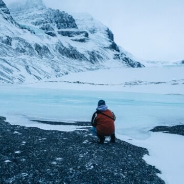 Persona sentada frente a un glaciar en retroceso, rodeada de nieve y montañas heladas.