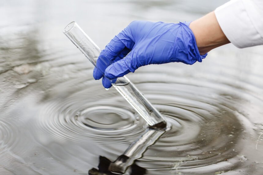 Mano con guante azul recolecta muestra de agua en tubo de ensayo desde un cuerpo acuático para análisis científico.