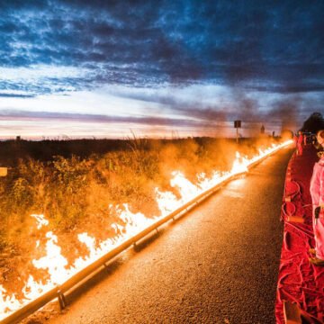 Protesta contra el uso de combustibles fósiles Activistas con trajes rojos observan una línea de fuego encendida en una carretera durante una protesta al atardecer, destacando el peligro de los combustibles fósiles.