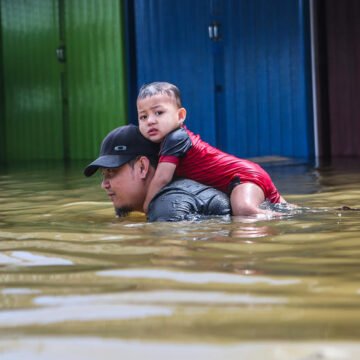 Rescate en medio de la inundación Hombre carga a un niño sobre su espalda mientras camina por una calle inundada con el agua hasta el pecho.