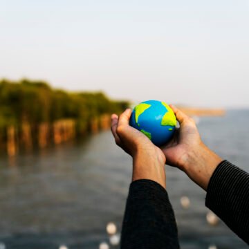 El planeta en nuestras manos Persona sosteniendo un pequeño globo terráqueo con ambas manos frente al mar y un paisaje natural al fondo.