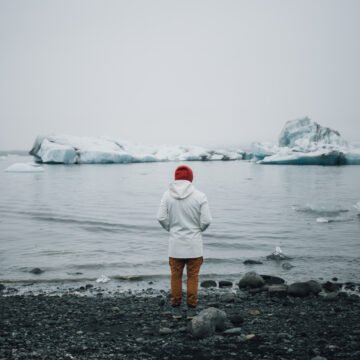 Reflexión ante el deshielo Persona con abrigo blanco y gorro rojo observa un paisaje polar con témpanos flotando sobre el agua en un ambiente frío y nublado.